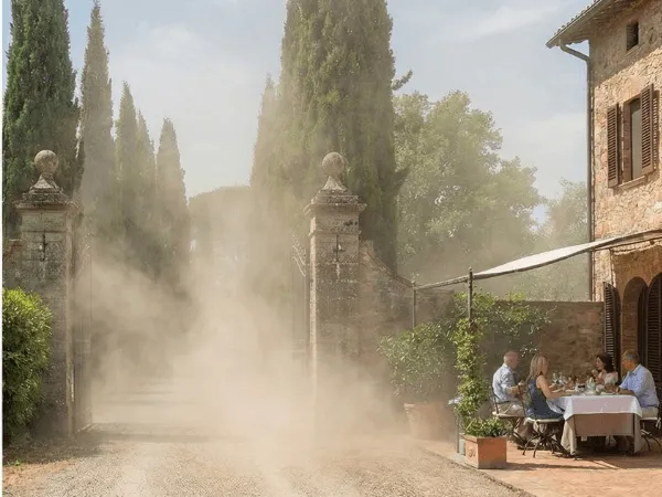 Un grande polverone si alza da una strada sterrata all'ingresso di una tenuta in pietra, investendo un tavolo dove un gruppo di persone sta pranzando all'aperto, visibilmente disturbato.