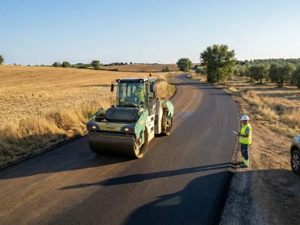 Un rullo compressore sta compattando il nuovo manto stradale nero appena steso su una strada di campagna ondulata
